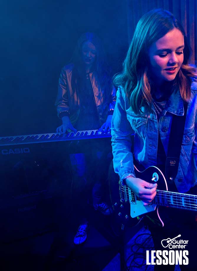 Teenager in blue jacket strumming electric guitar on stage.