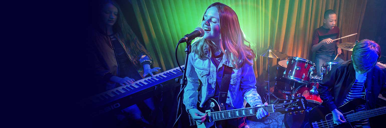 Teenager in blue jacket strumming electric guitar on stage with her band.